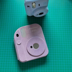 以下が含まれることがあります： 前面に「memory 2023」と印刷された薄い紫色のカメラケース。ケースはフェイクレザーの質感で、カメラレンズ用の円形の切り抜きがあります。