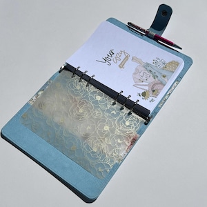 Puede incluir: Una agenda azul claro con un diseño floral en la portada. La agenda está abierta en una página en blanco con las palabras "Your Cozy" escritas en ella. Un bolígrafo está encima de la agenda.
