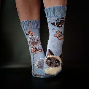 Peut inclure: Une paire de chaussettes en tricot bleu clair ornées d'un visage de chat siamois, de papillons marron et de fleurs de pissenlit. Les chaussettes présentent un bord-côte en haut et un motif intarsia détaillé dans des tons marron, crème et noir.