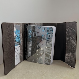 Puede incluir: Un planificador de cuero marrón con una cubierta transparente con la palabra "PLANNER". El planificador está abierto, revelando un cuaderno y un bolsillo con tarjetas. El interior tiene un diseño texturizado.