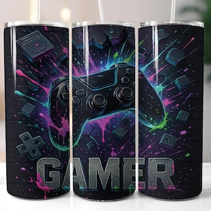 Puede incluir: Juego de tres vasos de acero inoxidable con temática de videojuegos. Cada vaso presenta un diseño de mando negro con salpicaduras rosas, moradas y verdes. La palabra "GAMER" se muestra en gris.