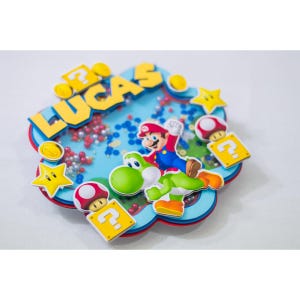 May include: A colorful, 3D cake topper featuring Mario and Yoshi from the Super Mario Bros. video game series. The topper is decorated with a blue, red, and yellow cloud shape, and the name "Lucas" is written in yellow letters. The topper is perfect for a Super Mario Bros. themed birthday party.