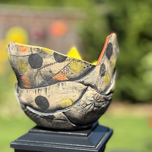 May include: A handmade ceramic bowl with a distinctive design. The bowl has a beige base with black, orange, and yellow patterns. The rim is orange. The bowl is on a black post.