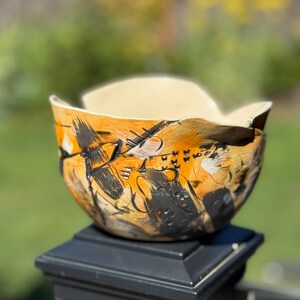 Puede incluir: Un cuenco de cerámica hecho a mano con un diseño abstracto único. El cuenco presenta una base naranja cálido con pinceladas negras y blancas. El borde tiene una forma ondulada y orgánica, y el interior es de color beige claro. El cuenco se exhibe sobre un poste negro.