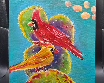 May include: Square canvas art featuring a vibrant painting of two cardinals perched on a cactus. The male cardinal is bright red, and the female is yellow. The background is a turquoise blue with peach-colored accents.