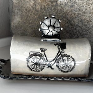 May include: A ceramic butter dish with a cream-colored lid featuring a black bicycle illustration. The lid has a silver-toned, wheel-shaped handle. The base is dark gray with a scalloped edge and small handles on each side.
