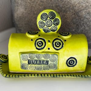 May include: A bright yellow ceramic butter dish with a rectangular base and a domed lid. The lid features a central handle and is decorated with black concentric circle patterns. The word BUTTER is embossed in black on the front panel.