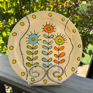 May include: A decorative ceramic plate with a floral design. The plate features three stylized flowers in orange, teal, and yellow, with black stems and leaves. The plate is cream-colored with yellow polka dots and is displayed on a silver metal stand.