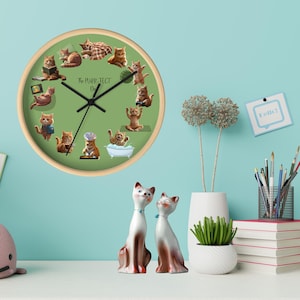 Op de afbeelding: Een ronde wandklok met een licht houten frame en een groene wijzerplaat met cartoonkatten in verschillende poses. De wijzers van de klok zijn zwart en de tekst "The PURR-FECT Day" staat op de wijzerplaat. Twee keramische kattenbeeldjes staan op de voorgrond.