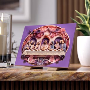 Op de afbeelding: Vierkante, paarse decoratieve tegel met een 3D-weergave van het Laatste Avondmaal. Het kunstwerk toont figuren die aan een tafel zitten, met bloemen- en vlinderaccenten. De tegel wordt op een houten standaard gepresenteerd.