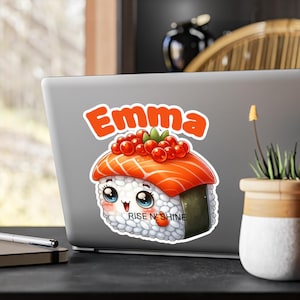 May include: A sticker featuring a cartoon sushi character with a salmon topping and red roe. The word "Emma" is above the sushi, and the phrase "RISE N' SHINE" is below. The sushi has large, expressive eyes and a smiling face.