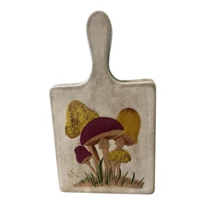 Puede incluir: Tabla de cortar de cerámica con asa, con un diseño de setas pintado a mano. Las setas tienen sombreros burdeos y amarillos con tallos marrones, sobre un fondo beige. Hierba verde pintada en la base.