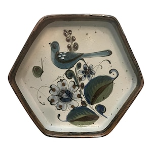 May include: Hexagonal ceramic plate with a hand-painted design. The plate features a blue bird perched on a floral branch with green leaves and white flowers. The rim is a dark brown color, and the background is off-white.