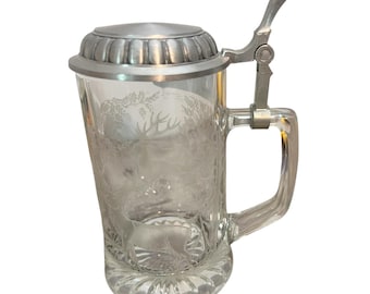 May include: Clear glass beer stein with a silver-colored metal lid and handle. The glass features an etched design of deer and trees. The stein has a hinged lid and a sturdy handle for easy gripping. A classic design for beer enthusiasts.
