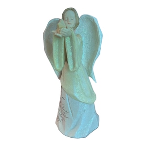 Può includere: Una statuina di angelo bianca con una finitura scintillante, che tiene un piccione nelle sue mani. L'angelo indossa un abito bianco con un bordo blu chiaro e ha un'aureola. La statuina ha un messaggio inciso sulla base.