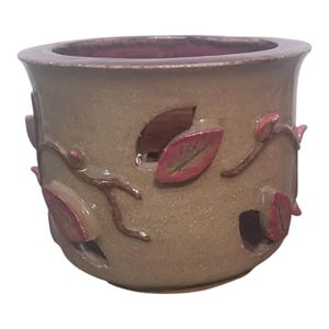 Puede incluir: Una maceta cilíndrica de cerámica con un exterior liso de color tostado que presenta motivos de hojas y enredaderas en relieve de color rosa oscuro. El recipiente incluye varias aberturas recortadas y un interior esmaltado en rosa oscuro.