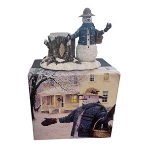 May include: A snowman figurine wearing a blue jacket, hat, and scarf, holding a basket, stands next to a tree stump. The figurine sits atop a box with a winter scene illustration featuring a house and another snowman.