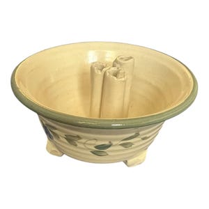 May include: A cream-colored ceramic planter with a green rim and floral design. The planter has three feet and a hole in the center for a plant.