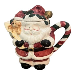 Könnte beinhalten: Eine Keramik-Teekanne in Form des Weihnachtsmanns mit rotem Hut, Mantel und gestreiftem Griff. Die Teekanne hat einen weißen Bart, schwarze Handschuhe und eine goldene Gürtelschnalle. Der Weihnachtsmann hält eine Liste mit einem Teddybären.