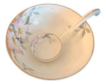 May include: A white ceramic serving bowl with a matching ladle. The bowl has a floral design with pink flowers and green leaves. The bowl and ladle have a gold trim.
