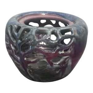 Puede incluir: Un pequeño recipiente de cerámica redondo con un intrincado diseño de celosía calada que recuerda a las ramas de los árboles. La superficie tiene un esmalte brillante multicolor con tonos mezclados de azul profundo, morado y gris.