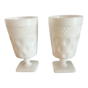 May include: Two white, vintage-style goblets. The glasses have a square base, a short stem, and a textured design. The upper portion of the glass features a diamond pattern, while the lower portion has a series of raised circles.