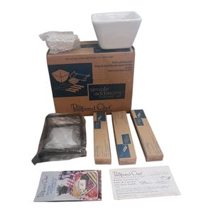 May include: A white square fondue bowl sits atop a cardboard box with the text "Simple Additions". The box contains fondue forks and other accessories. Additional boxes and a black carrying case are also visible.