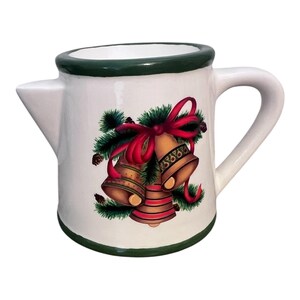 Puede incluir: Jarra de cerámica blanca con borde y asa verdes. La jarra presenta un diseño festivo de campanas doradas, un lazo rojo y agujas de pino verdes, ideal para la decoración navideña.