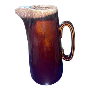 May include: A vintage ceramic pitcher with a glossy finish. The pitcher features a dark brown exterior with a lighter brown and tan rim. It has a curved handle and a spout for pouring. The pitcher is likely used for serving beverages.