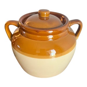 Könnte beinhalten: Ein brauner und beiger Keramik-Bohnentopf mit Deckel. Der Topf hat zwei Griffe und eine abgerundete Form.