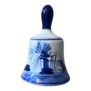 May include: A ceramic bell with a dark blue handle and a white body. The body is decorated with a blue windmill design. The bell has a flared base and a rounded top, with blue accents around the edges.
