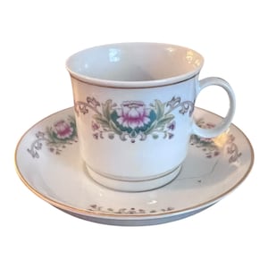 May include: A white porcelain teacup and saucer set with a floral design. The cup and saucer feature a gold trim and a pink and green floral pattern.