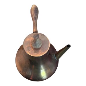 Pode incluir: Uma chaleira de latão com uma pega e tampa de madeira. A chaleira tem um corpo arredondado e um bico lateral.