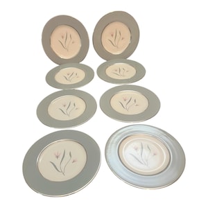 May include: A set of eight decorative plates with a floral design. The plates have a light gray border and a cream-colored center with a delicate floral illustration in pink and green. The plates vary in size.