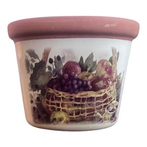 May include: A white ceramic planter with a pink rim features a painted image of a basket overflowing with fruit, including grapes, apples, and pears. The basket is woven with a brown and tan pattern.