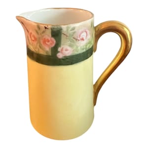 May include: A small, antique cream pitcher with a pale yellow body and a gold-colored handle. The top section features a green band with pink rose floral detailing. The pitcher has a spout for easy pouring.