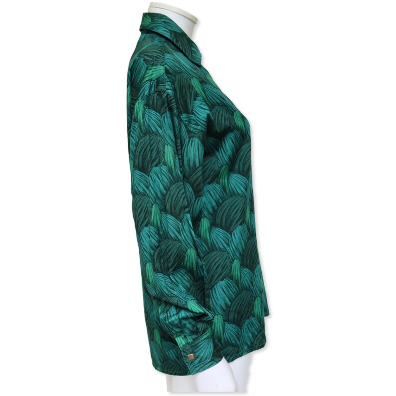 May include: A long-sleeved shirt with a green leaf pattern. The shirt is displayed on a white mannequin. The fabric has a repeating pattern of dark and light green leaves. The cuffs have a button closure. The collar is folded down.