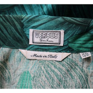 May include: Close-up of a green and teal patterned fabric with a VERSUS Gianni Versace label. The label is white with black text and a black border. A tag reads "Made in Italy".