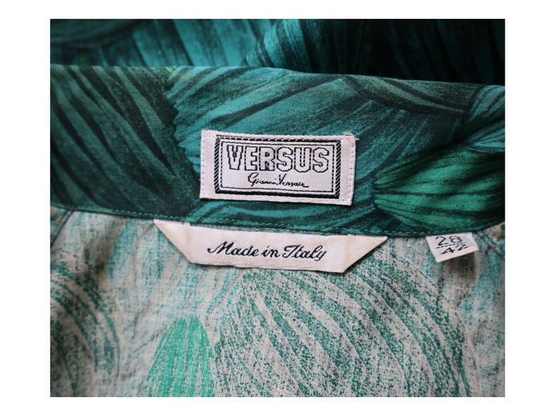May include: Close-up of a green and teal patterned fabric with a VERSUS Gianni Versace label. The label is white with black text and a black border. A tag reads "Made in Italy".