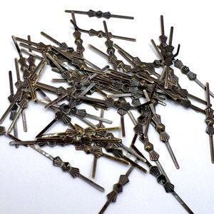 Może przedstawiać: Duża sterta antycznych, złotych metalowych klamer w kształcie kokardki. Klamry mają około 2,5 cm długości i posiadają ozdobny wzór.