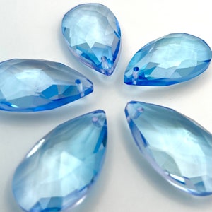 May include: Five light blue, faceted teardrop-shaped beads. Each bead has a hole at the top for stringing. The beads are arranged in a circular pattern, showcasing their translucent quality and geometric design.