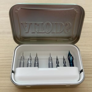 May include: A set of calligraphy pen nibs in a metal tin. The tin is open, revealing several silver nibs and one blue nib. The tin is light green with the word "VILOIDS" embossed on the inside of the lid.