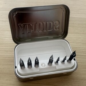 May include: A rectangular metal tin, open to reveal a white tray holding seven black pen nibs. The lid of the tin has the word "VILOTID2" embossed. The tin is on a light wooden surface.
