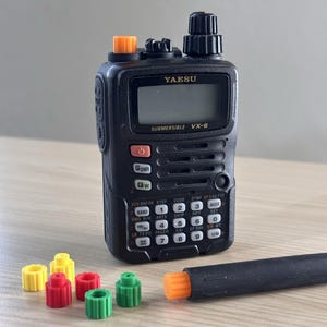May include: Black Yaesu VX-6 submersible radio with an orange top and a black antenna. Several colored knobs in yellow, red, and green are next to the radio. The radio has a screen and multiple buttons.