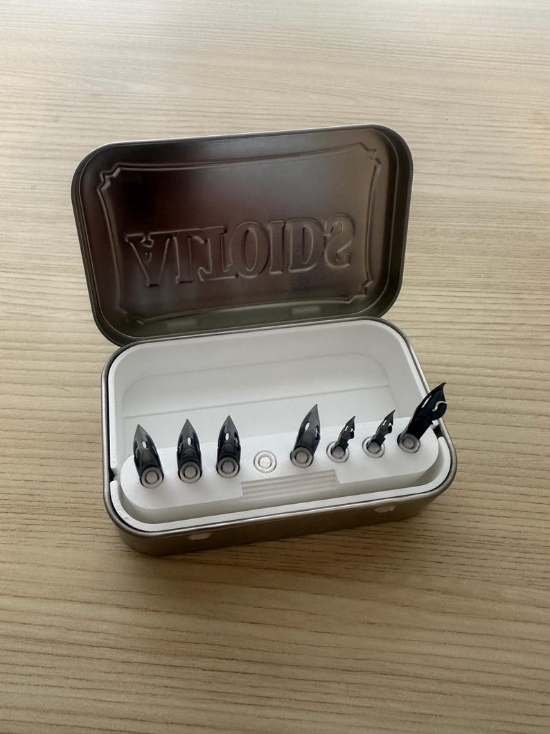 May include: A rectangular metal tin, open to reveal a white tray holding seven black pen nibs. The lid of the tin has the word "VILOTID2" embossed. The tin is on a light wooden surface.
