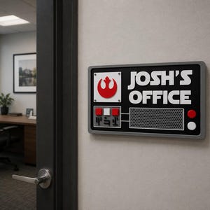 May include: A rectangular office sign with a black background and grey border is mounted on a wall. The sign features the text "JOSH'S OFFICE" in white, along with a red and white symbol. The sign has a retro, sci-fi aesthetic.