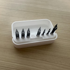 May include: A white rectangular container holding several black pen nibs of varying sizes. The nibs are arranged in a row, with the largest on the left and the smallest on the right. The container is on a light brown wooden surface.