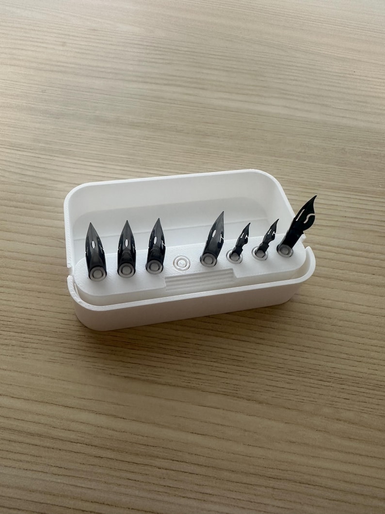 May include: A white rectangular container holding several black pen nibs of varying sizes. The nibs are arranged in a row, with the largest on the left and the smallest on the right. The container is on a light brown wooden surface.