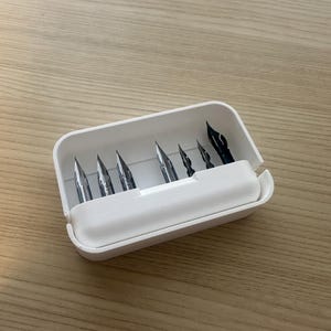 May include: A white plastic pen nib storage case containing several metal pen nibs. The nibs are of various sizes and shapes, with a few black nibs visible. The case is open, revealing the contents, and sits on a light brown wooden surface.