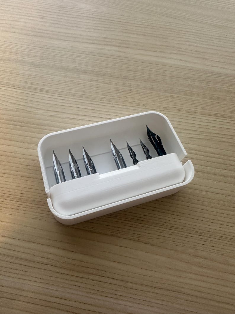 May include: A white plastic pen nib storage case containing several metal pen nibs. The nibs are of various sizes and shapes, with a few black nibs visible. The case is open, revealing the contents, and sits on a light brown wooden surface.